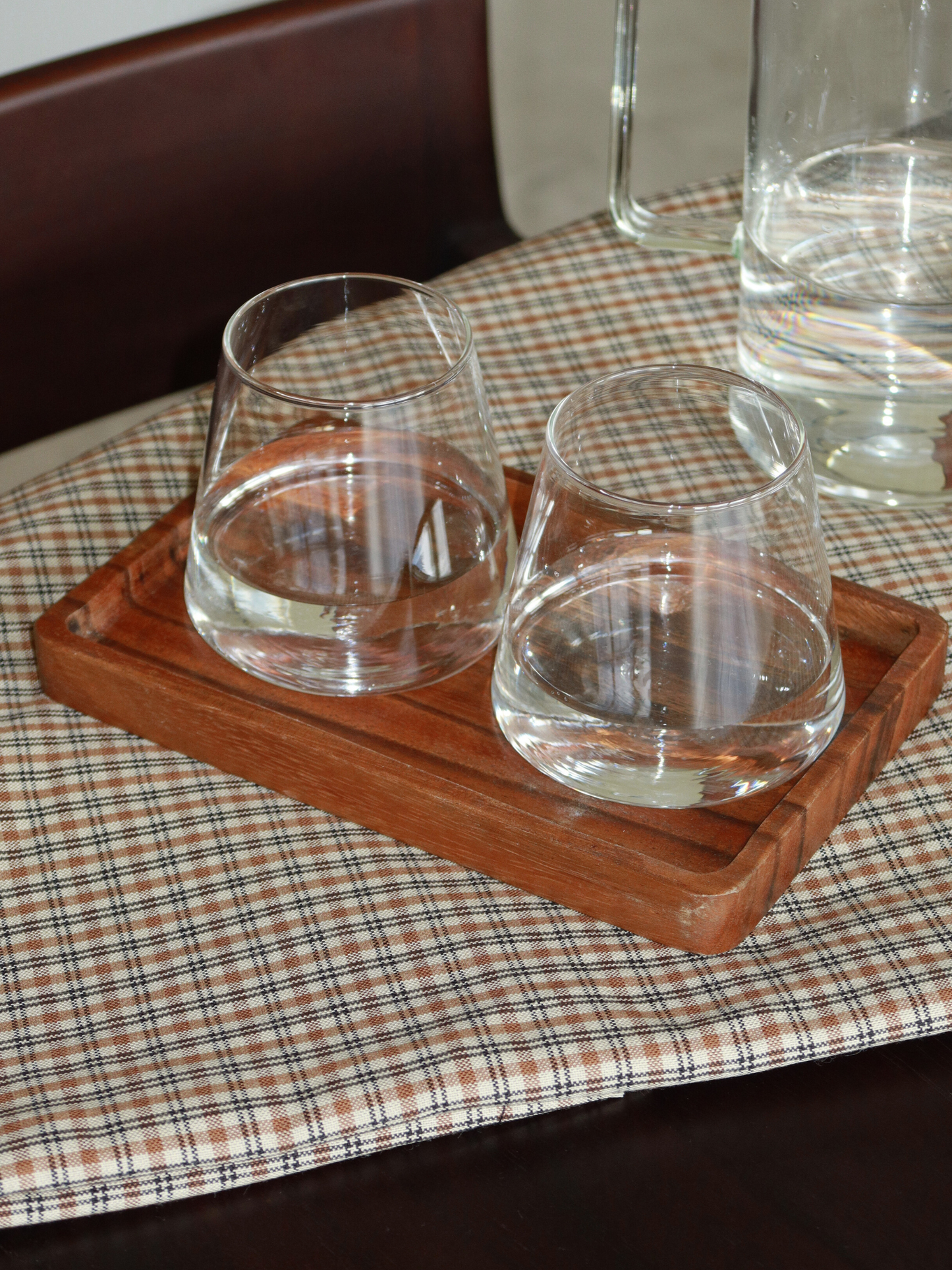 Plateau de service rectangulaire en bois, dans une teinte brun chaud et profond, posé sur une nappe à carreaux neutre aux tons beige et brun doux. Le plateau présente des bords légèrement surélevés et un design lisse et minimaliste, accueillant deux verres à eau transparents sans pied, partiellement remplis d’eau. En arrière-plan, une carafe en verre et une chaise en bois foncé renforcent l’esthétique chaleureuse et naturelle. L’ensemble dégage une atmosphère calme, simple et soigneusement mise en scène — r