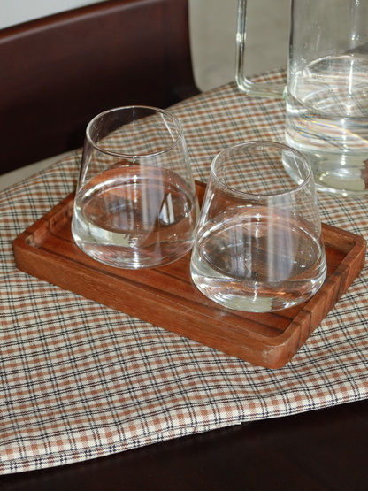 Plateau de service rectangulaire en bois, dans une teinte brun chaud et profond, posé sur une nappe à carreaux neutre aux tons beige et brun doux. Le plateau présente des bords légèrement surélevés et un design lisse et minimaliste, accueillant deux verres à eau transparents sans pied, partiellement remplis d’eau. En arrière-plan, une carafe en verre et une chaise en bois foncé renforcent l’esthétique chaleureuse et naturelle. L’ensemble dégage une atmosphère calme, simple et soigneusement mise en scène — r